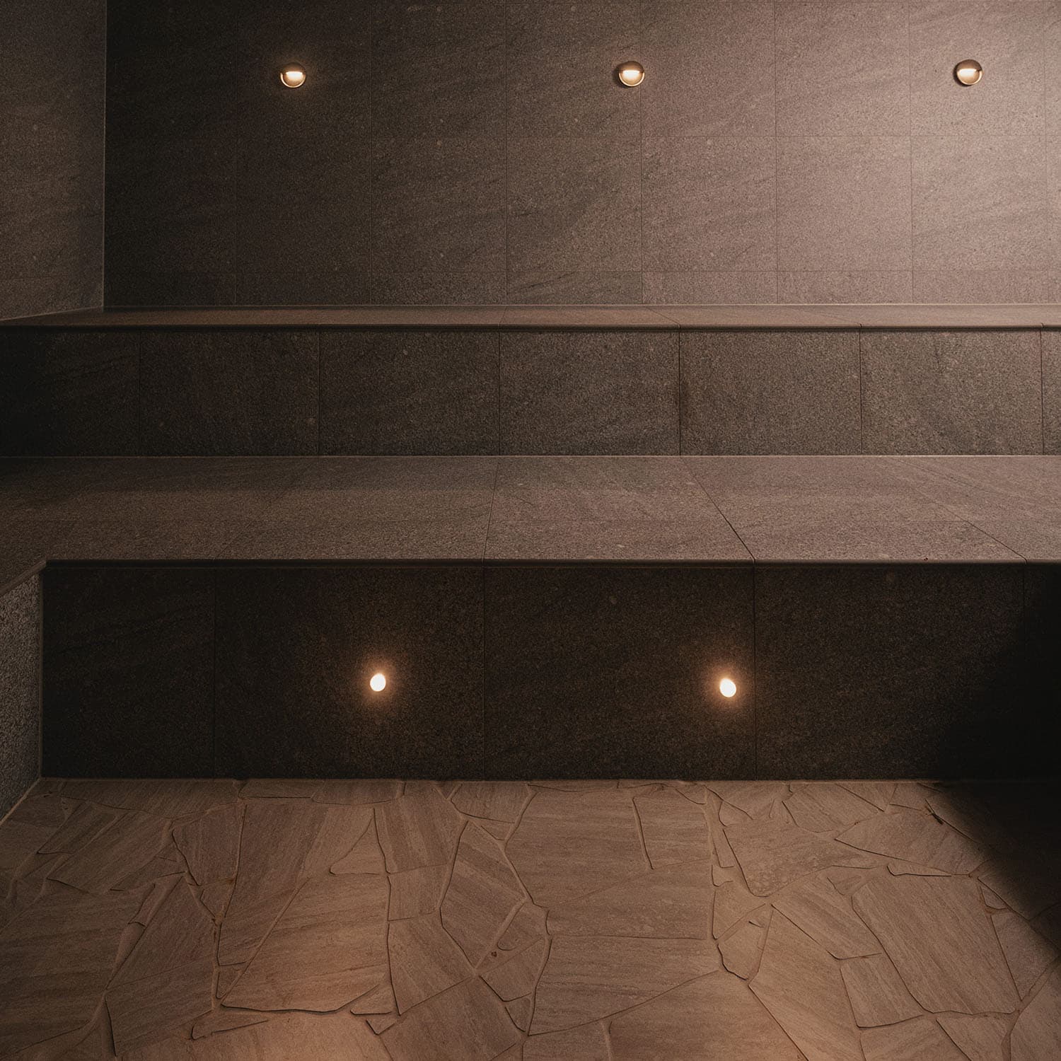 An empty steam room in dark stone, with a bench running along the wall and small recessed lights set into the wall above and below it