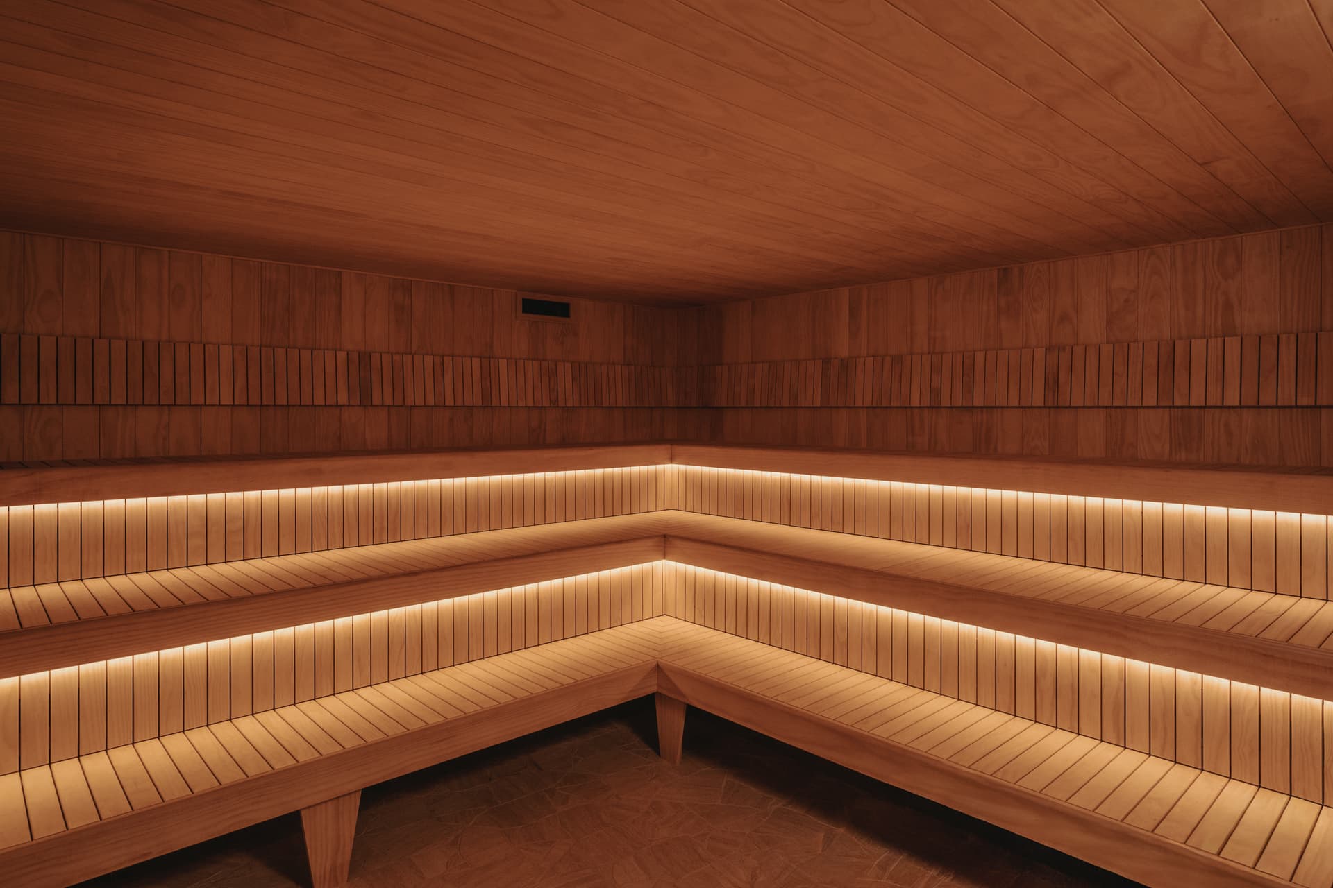 Empty cedar-lined Finnish sauna with two tiers of slatted bench seating wrapping the corner and warm strip lighting beneath each bench