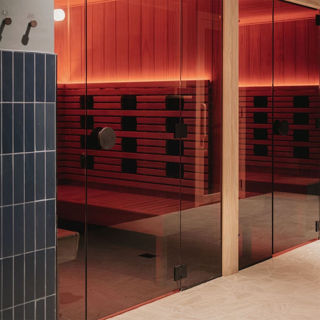 Two infrared Saunas behind glass doors, their timber benches lit red, beside a tiled wall of numbered towel hooks