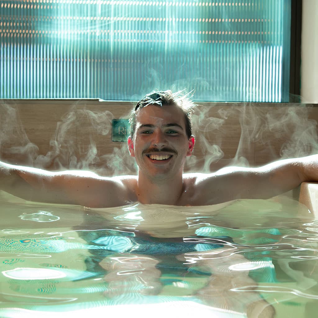 A smiling man rests his arms along the edge of a cold plunge pool, steam rising from the water around him