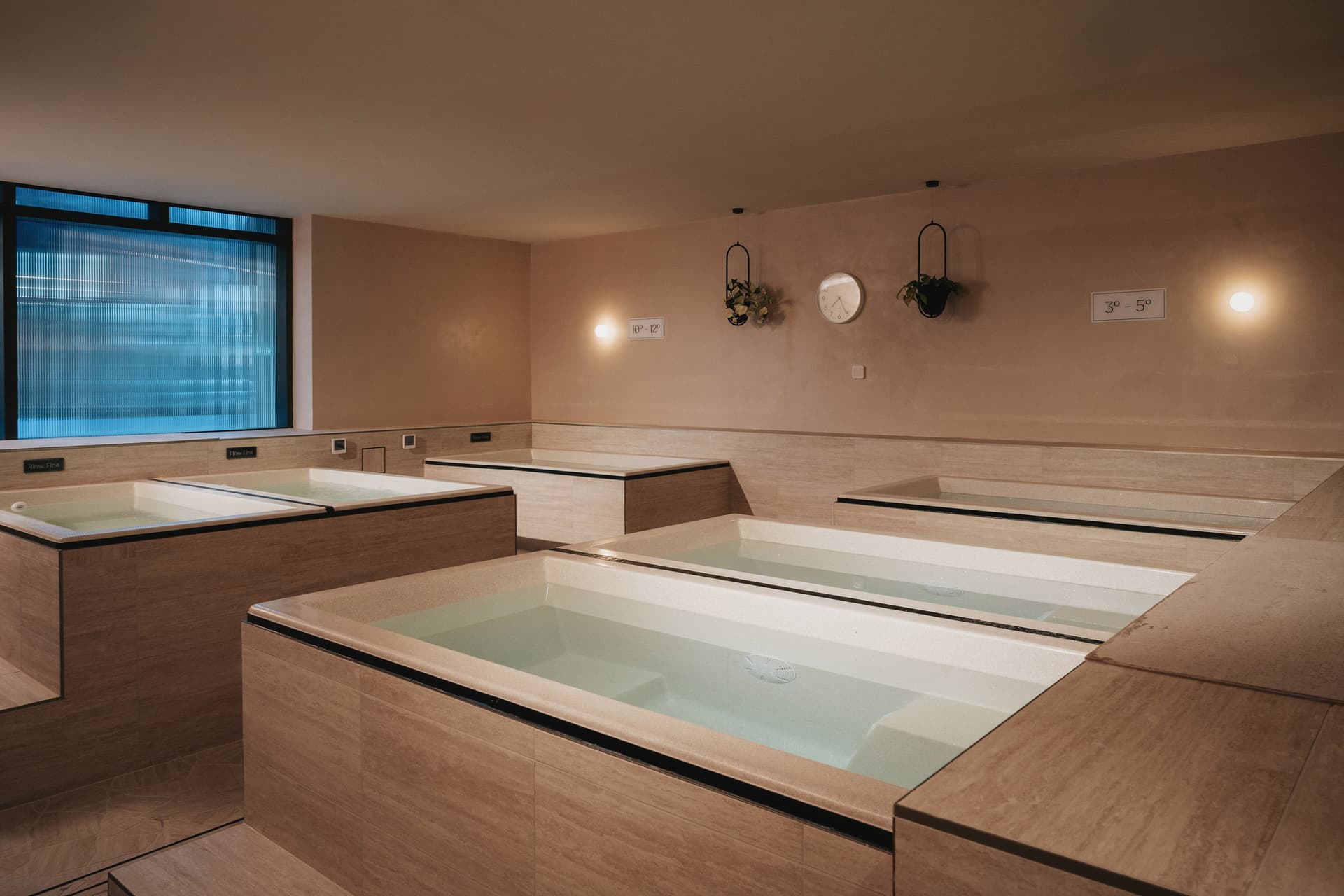 Room of four stone-clad cold plunge baths with temperature signage on the wall and a fluted glass window