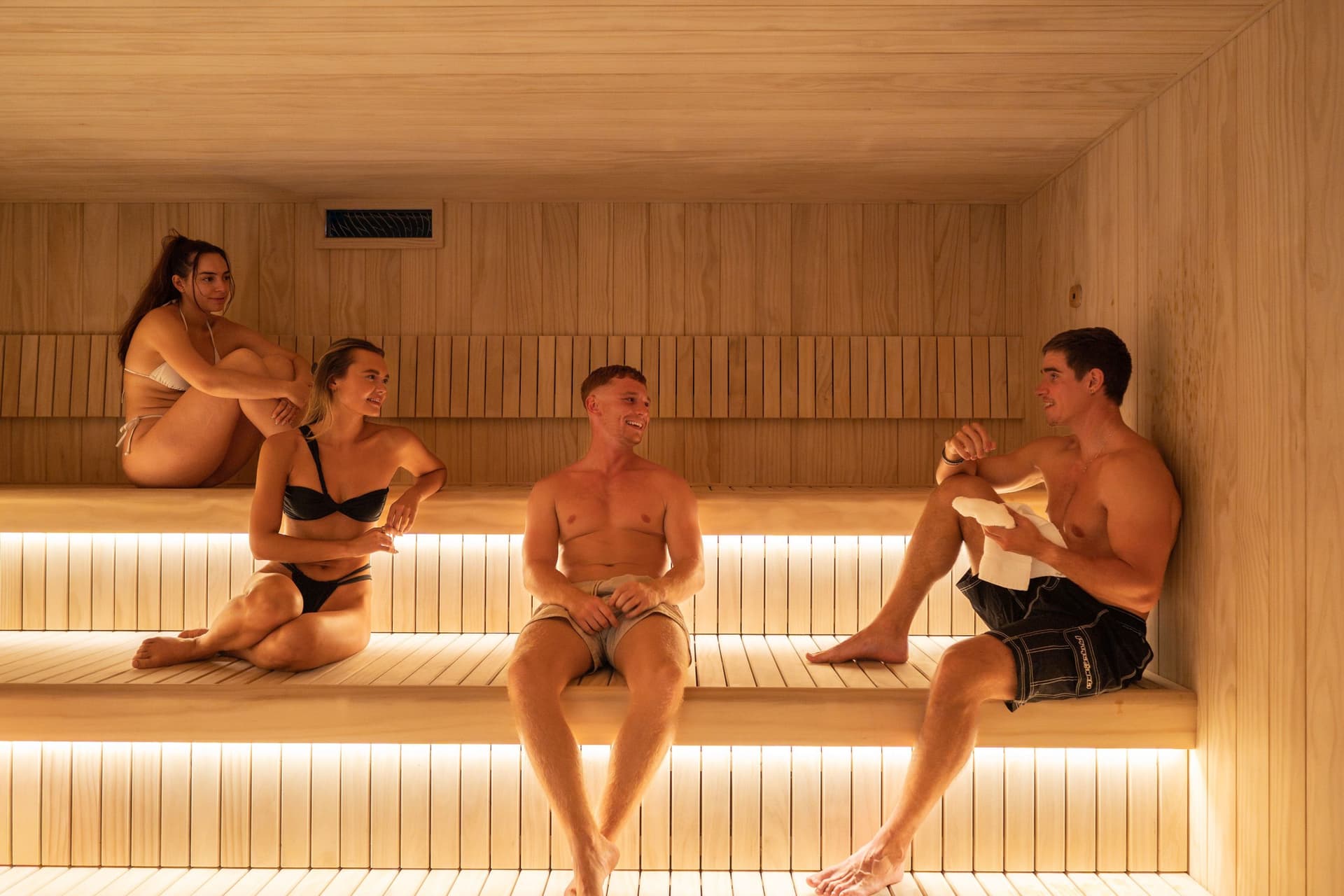 Four friends spread across the tiered benches of the sauna, talking