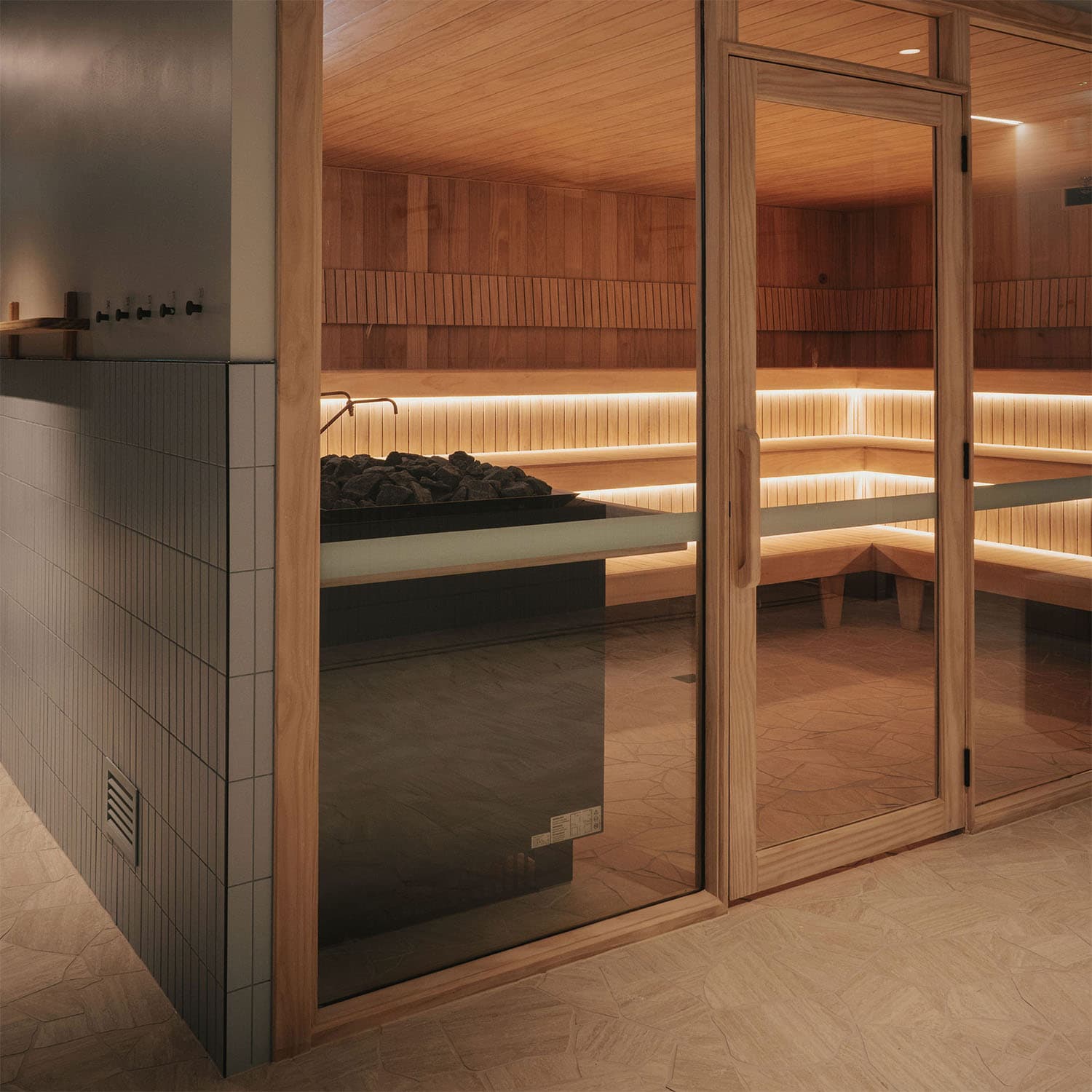 A glazed sauna cabin seen from the changing area, its tiered timber benches lit by a strip of warm light beneath the seating
