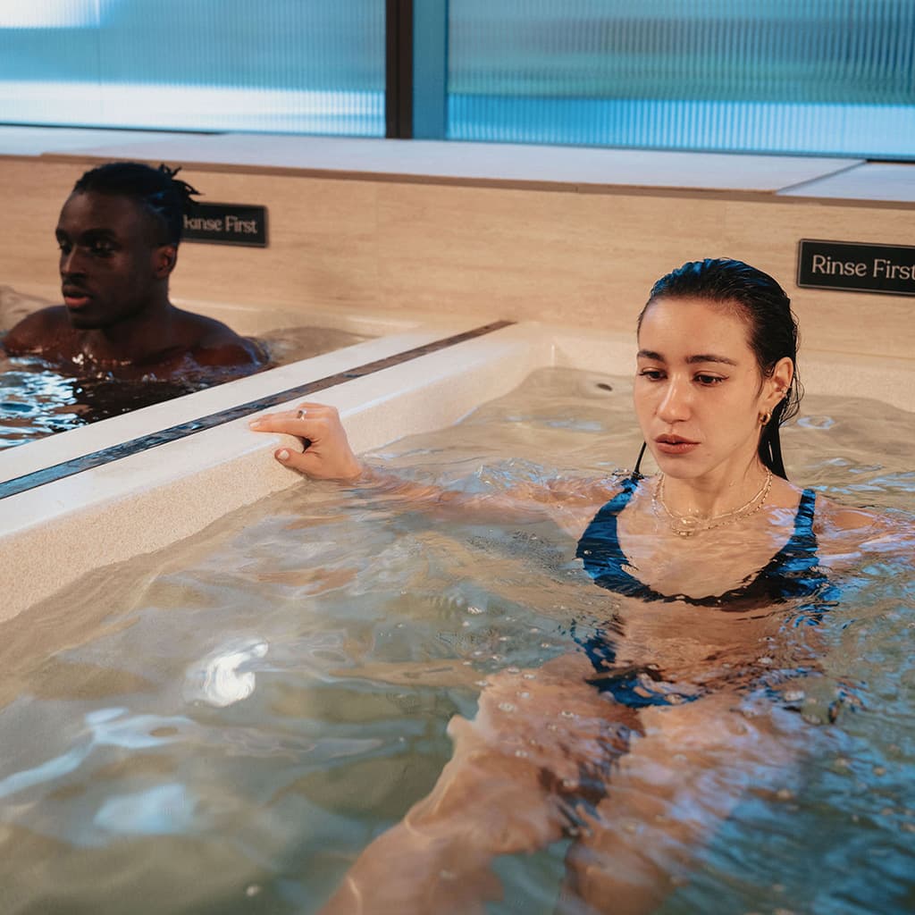 A woman in a blue swimsuit stands in one cold plunge pool talking to a man in the pool beside her, both chest-deep in the water