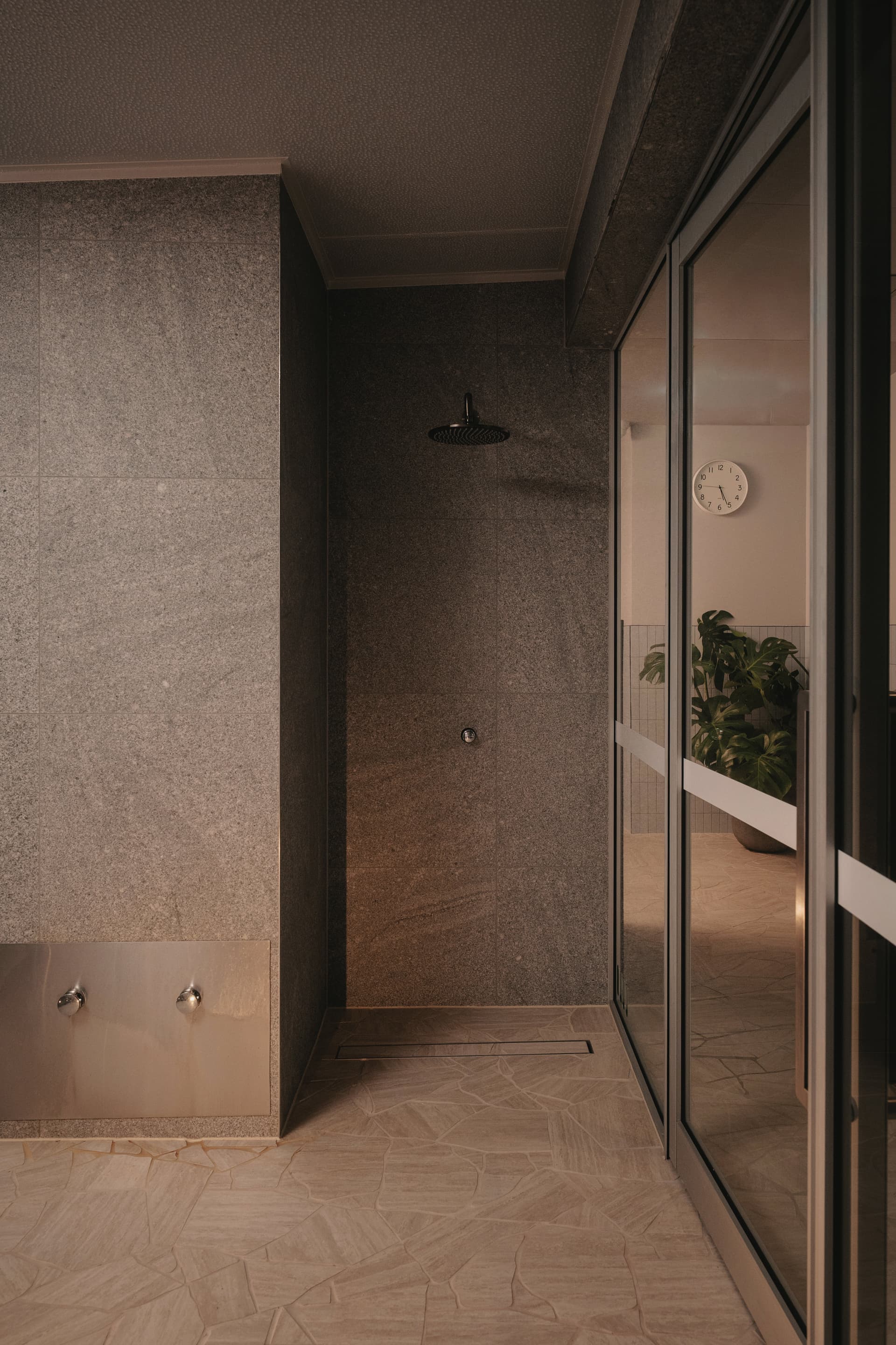 Open shower alcove tiled in dark stone with a black rain shower head and brushed metal wall fittings, beside a glazed wall onto a planted lounge
