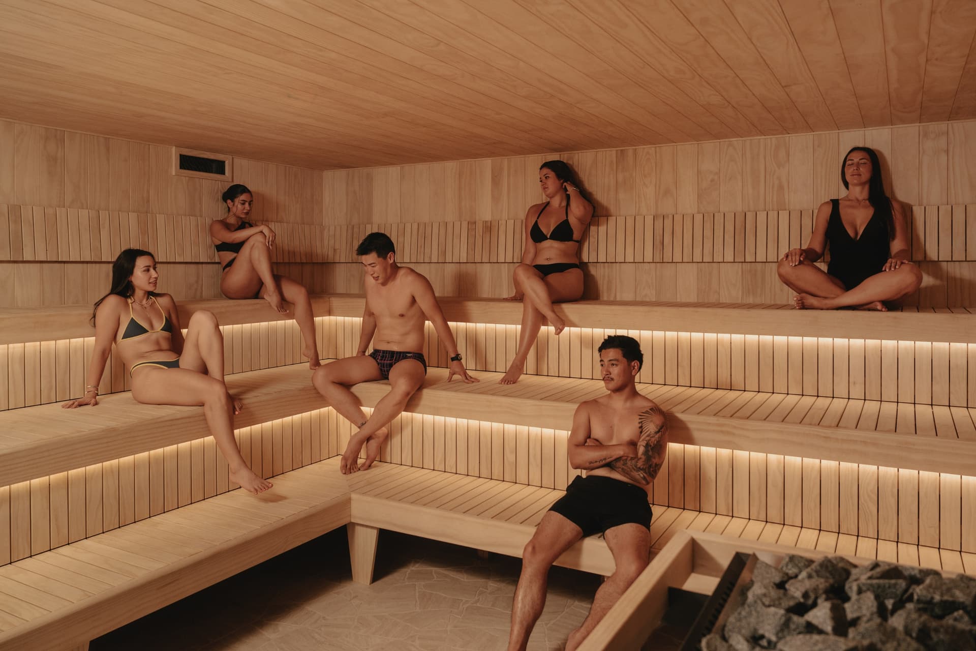 Six people using the tiered benches of a large Finnish sauna, the stove and stones in the foreground