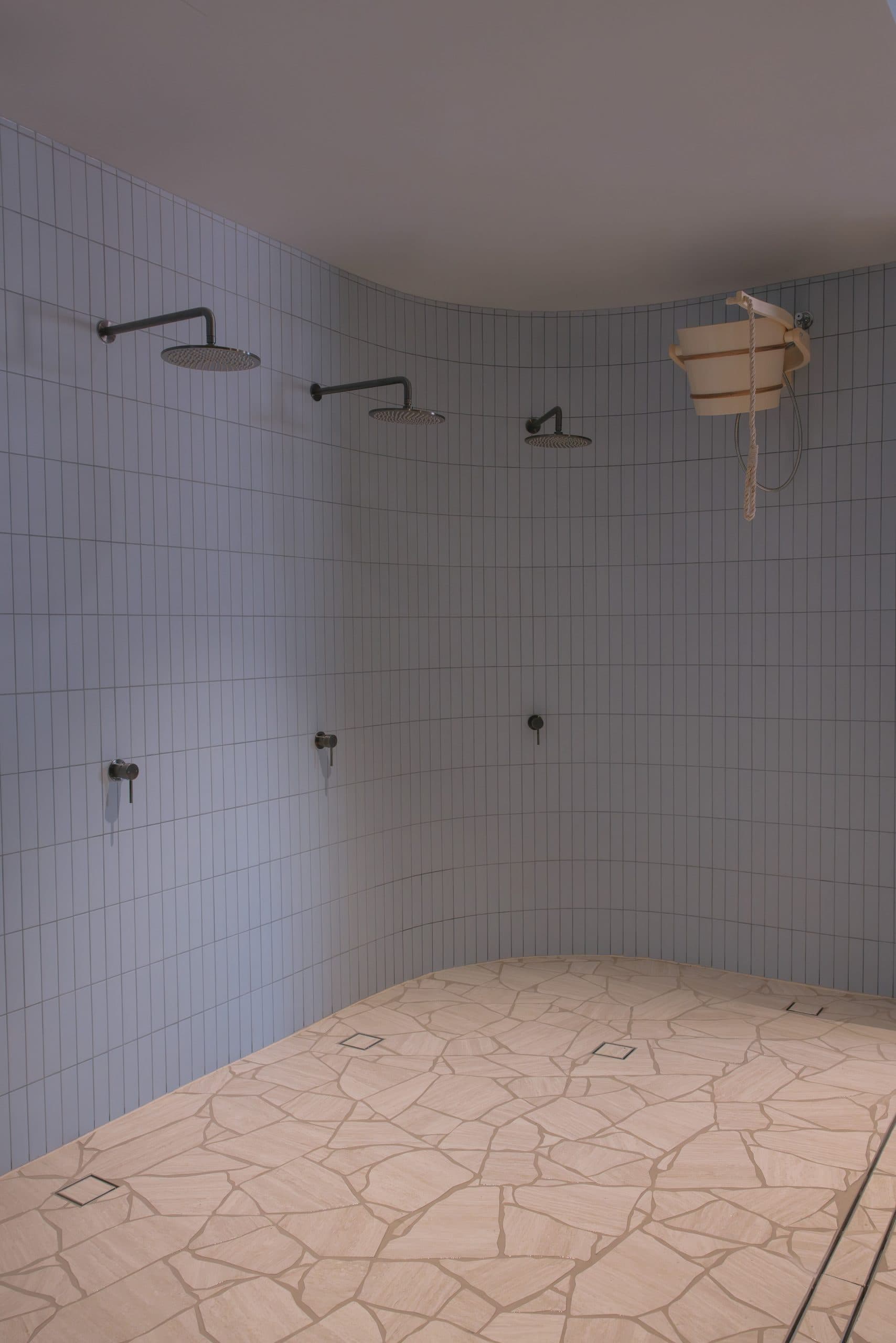 A white-tiled communal shower area with a bucket shower and terrazzo floor