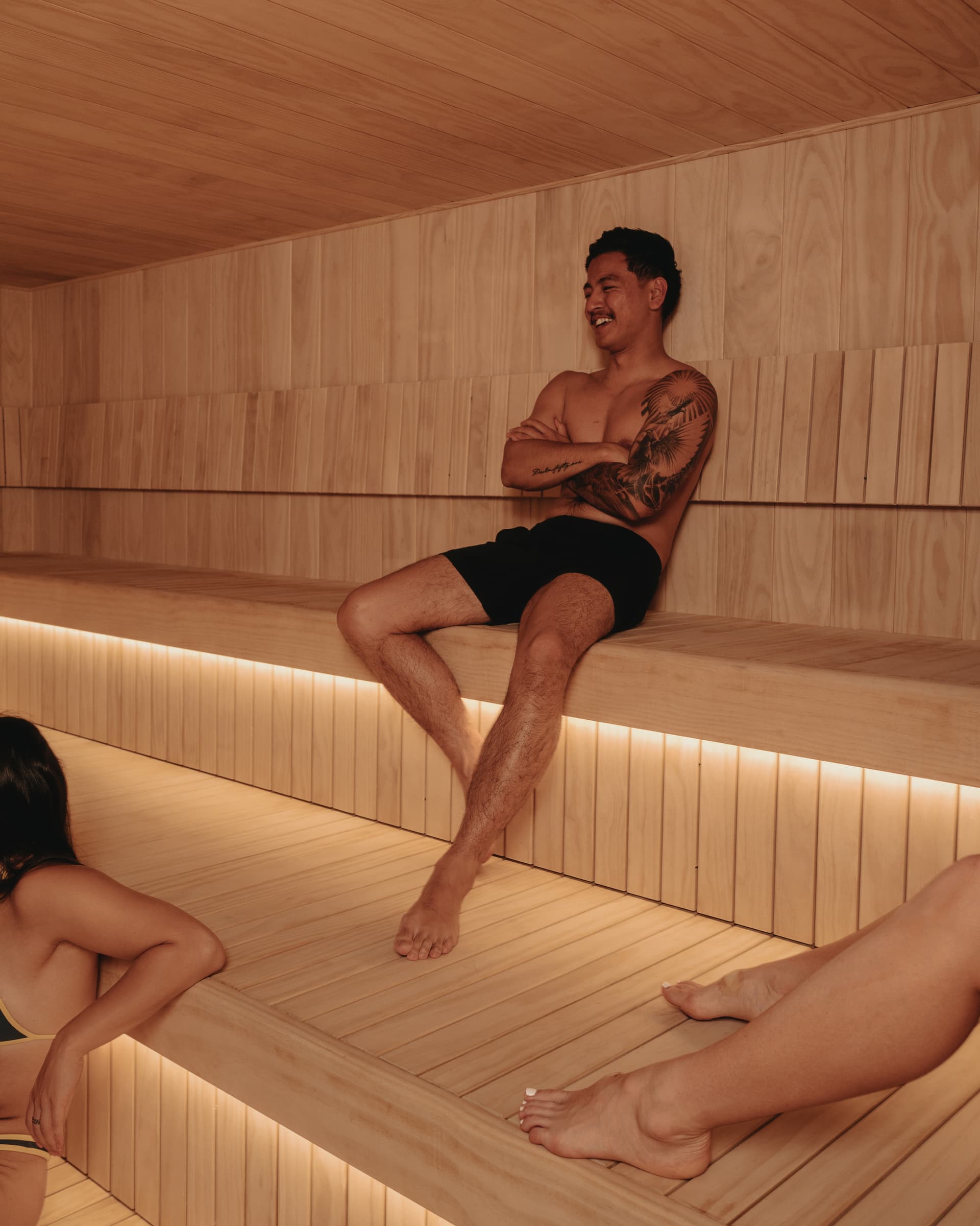 Three people sitting along the bench of a timber-lined Finnish sauna, talking and gesturing mid-conversation