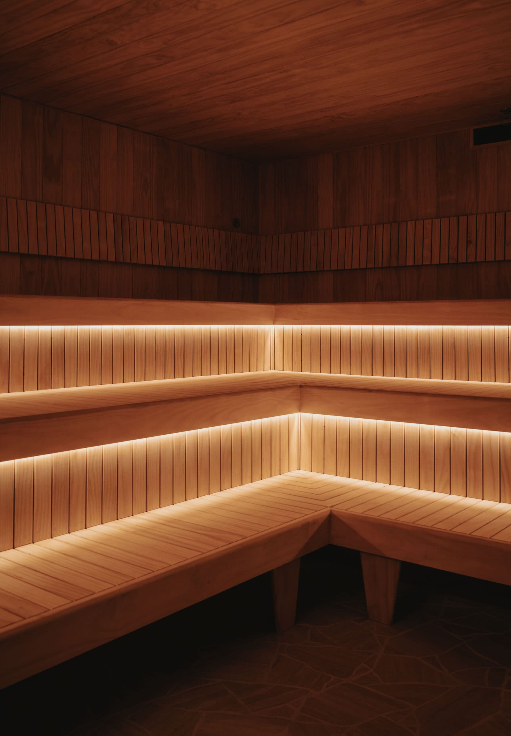 The corner of an empty sauna, its tiered benches wrapping around two walls and lit from beneath by warm strip lighting