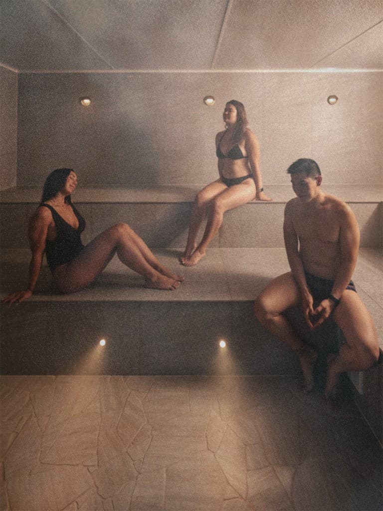 Three people sitting on the tiered stone benches of a steam room, steam hanging in the air under round wall lights