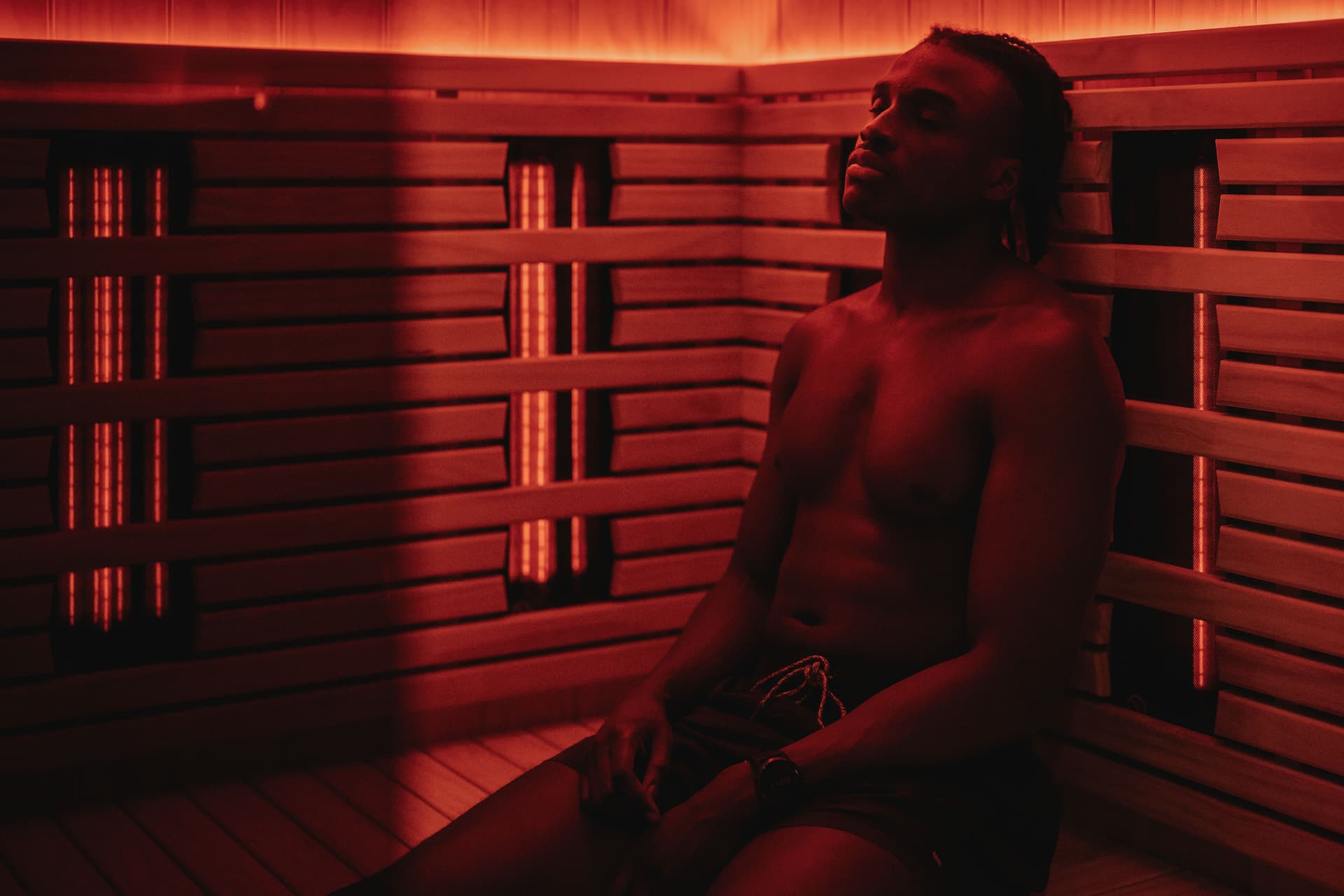 A person leaning back with eyes closed in the corner of an infrared sauna, heater elements glowing along the slatted walls