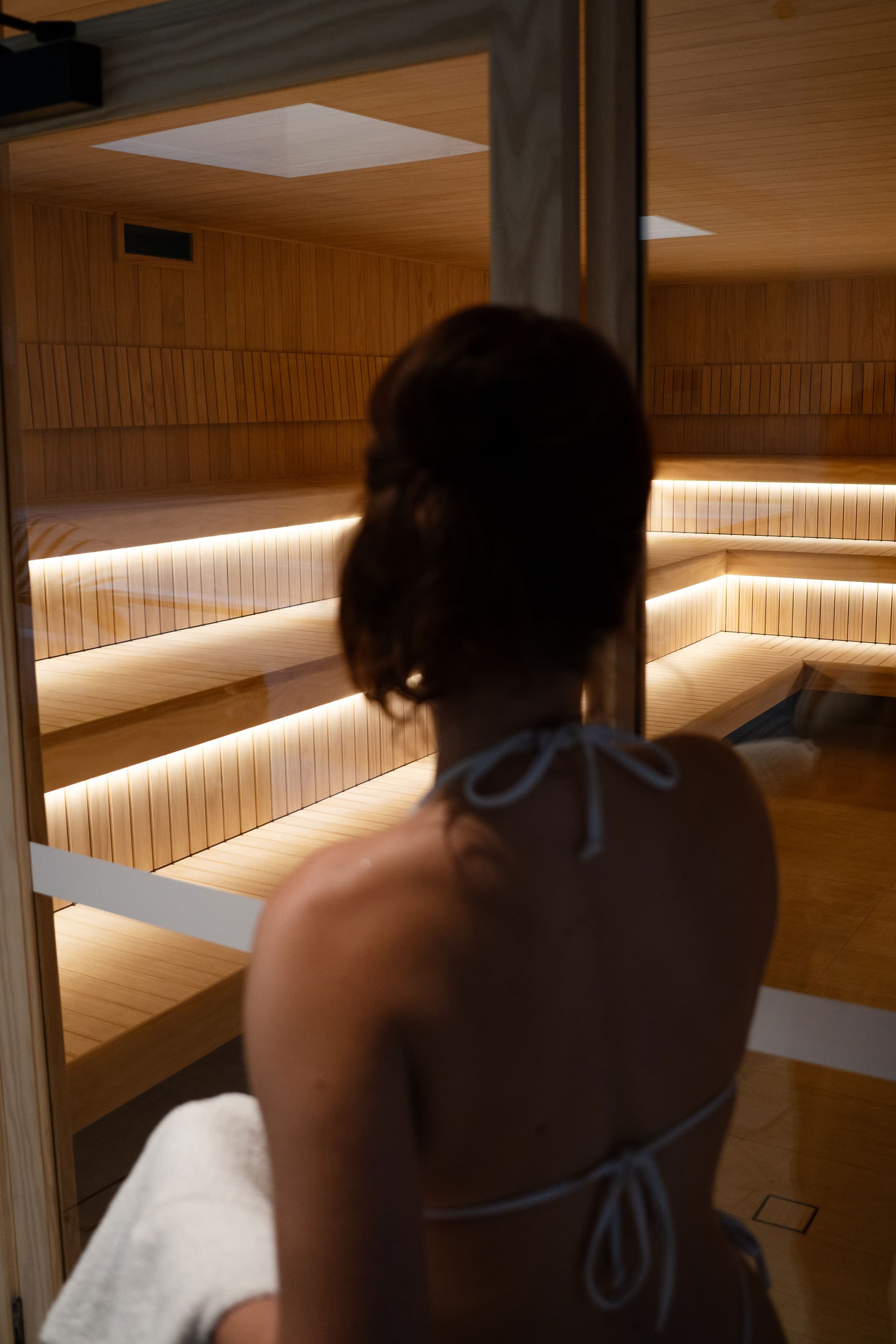 Person seen from behind at the glass door of a Finnish sauna, looking in at strip-lit tiered timber benches