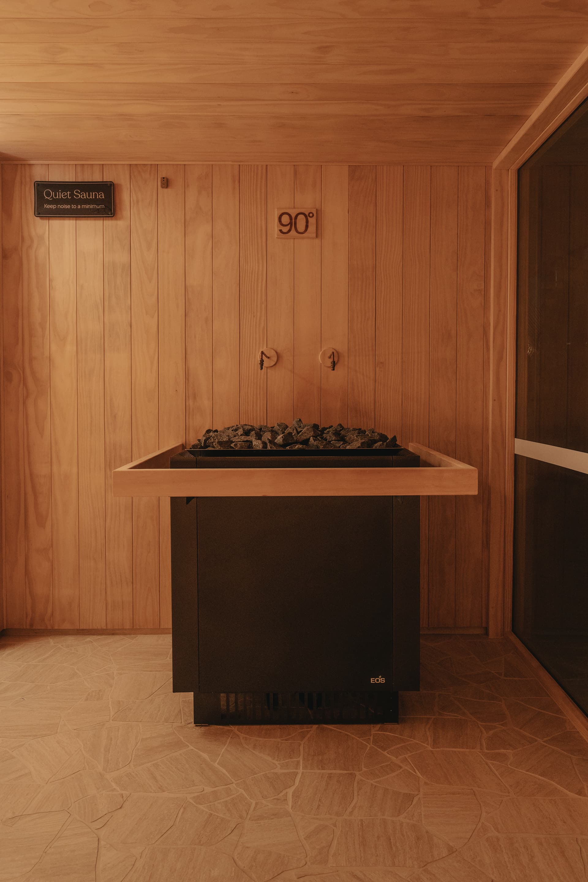 Black stone-filled sauna heater against timber wall panelling beneath a quiet-sauna sign and a ninety degree temperature plaque
