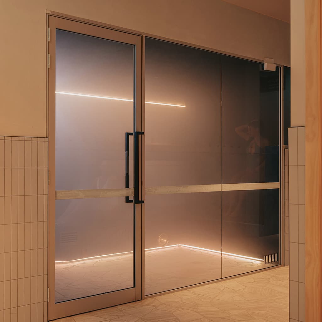 The glass doors of a steam room seen from the corridor outside, warm light spilling from within